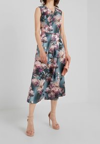 Blommig midiklänning med en teal bakgrund, ärmlös design, plisserad kjol och en nyckelhålsringning. Kombinerad med nakna remsandaler och en rosa clutch.