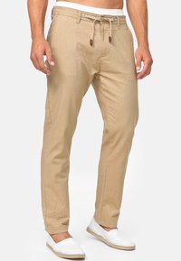 Pantaloni in cotone beige con una vestibilità rilassata, dotati di una vita elastica con coulisse e dettagli in legno. Indossati con scarpe bianche slip-on.