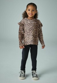 Leopardenmuster Langarmtop mit Rüschenschultern, kombiniert mit schwarzen Leggings und schwarzen Turnschuhen mit weißen Schnürsenkeln und strukturiierter Sohle.