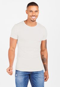 Uomo sorridente che indossa una maglietta bianca semplice e jeans blu, in piedi rivolto verso l'osservatore, con tatuaggi visibili sulle braccia, su uno sfondo bianco.