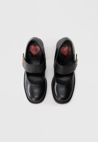 Sapatos de couro pretos com ponta redonda e tira no tornozelo. Apresentam detalhes em metal dourado e o texto "Love Moschino" em um palmilha em formato de coração vermelho.