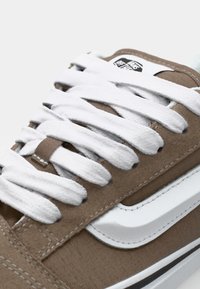 Braune Leinenschuhe mit weißen Schnürsenkeln, ausgestattet mit einem weißen Lederstreifen an der Seite und weißen Nähdetails, entworfen für den Freizeitgebrauch.