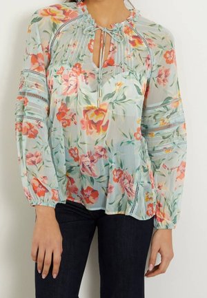 Jasnoniebieska, kwiatowa bluzka z długim rękawem, wykonana z przezroczystej tkaniny, z dekoltem na kokardkę i wstawkami koronkowymi, noszona z ciemnymi spodniami.