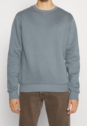 Homme portant un sweat-shirt bleu uni délavé à col rond avec poignets et ourlet côtelés, associé à un pantalon marron, debout sur un fond blanc.