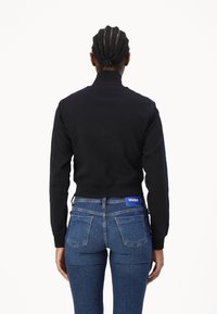 Schwarzes, kurz geschnittenes Sweatshirt mit hohem Kragen, gerippten Bündchen und Seitenstreifen. Kombiniert mit blauen Jeans aus Denim, die ein gebrandetes Rückenpatch aufweisen.