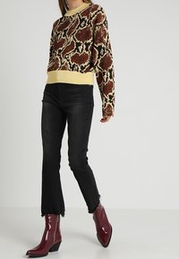 Strickpullover mit einem braunen und beigen Schlangenmuster, kombiniert mit schwarzen Jeans mit fransigem Saum und glänzenden roten Stiefeletten.