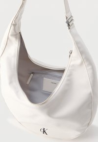 Λευκή ώμου τσάντα Calvin Klein με ασημί φερμουάρ, ρυθμιζόμενο λουράκι, εσωτερική τσέπη με φερμουάρ και μαύρο λογότυπο "cK" μπροστά.