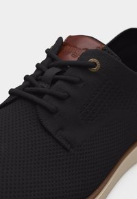 Μαύρο πλεκτό casual παπούτσι με καφέ δερμάτινο ταμπελάκι στη γλώσσα, μαύρα κορδόνια και μπεζ σόλα, που δείχνει λεπτομερή υφή στο πάνω μέρος και τις τρύπες για τα κορδόνια.