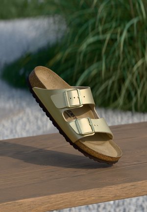 Guld to-strop sandal med metalspænder og kork-fodseng, der hviler på en træoverflade udendørs med en sløret grøn baggrund.