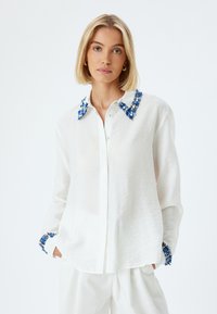 Camisa de color blanco con adornos florales azules en el cuello y los puños, con un tejido texturado y un diseño de corte holgado.