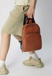 Sac à dos en cuir marron avec double fermeture éclair, poignée supérieure et bretelles noires rembourrées. Texture lisse et forme rectangulaire compacte.