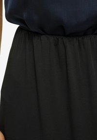 Robe noire présentant un corsage lisse et fluide avec une jupe plissée. Le tissu a un léger éclat, et la taille est froncée pour ajouter de la texture.