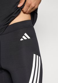 Schwarze Sportleggings mit glatter Textur, akzentuiert durch drei weiße diagonale Streifen und ein weißes Logo an der Seite.