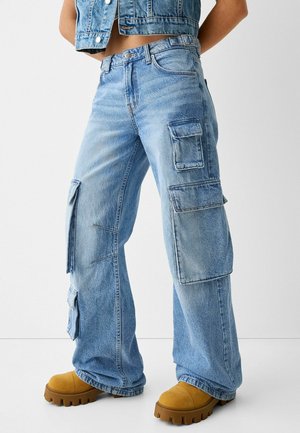 Osoba ubrana w jasnoniebieskie szerokie spodnie typu cargo z licznymi dużymi kieszeniami oraz brązowe buty na masywnej podeszwie na jednolitym tle.