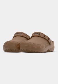 Paire de Crocs marron sans lacets avec brides de talon réglables et semelles texturées sur fond blanc uni.