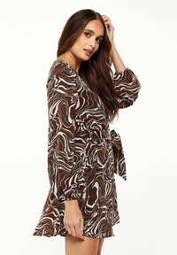 Brown patterned dress with swirling white designs, featuring long sleeves, a tied waist, and a flared skirt. Smooth texture.