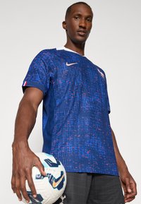 Tricou de fotbal Nike de culoare albastru marin cu un model texturat, guler alb și accente roșii. Modelul ține o minge de fotbal albă cu detalii albastre și negre.