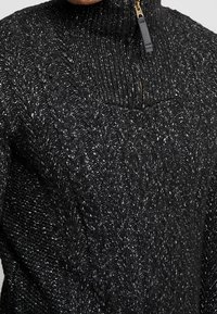 Pull noir à col montant avec un motif en maille torsadée texturée et un col zippé, présentant un mélange de fibres foncées et claires pour plus de profondeur.