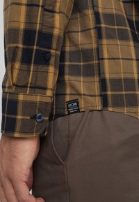 Brown and navy plaid shirt with long sleeves, button details, and a KIOMI brand tag, paired with beige pants. Soft fabric texture visible.