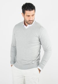 Un pull léger gris clair en v, en tissu doux, avec des manches longues et des poignets côtelés, associé à une chemise blanche à col et à un pantalon crème.