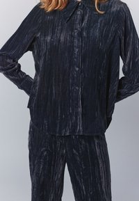 Sötét tengerészkék gombos ing, hegyes gallérral, bársonyos textúrával és függőleges csíkokkal; hozzá illő nadrággal párosítva.