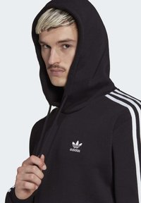 Black hoodie with a white Adidas logo on the chest, featuring white three-stripe accents on the sleeves and a drawstring hood.