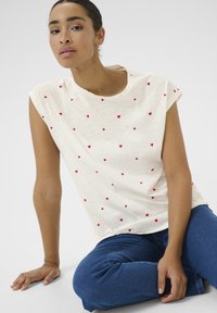Chemise blanche à manches courtes avec de petits motifs de cœurs rouges dispersés. Texture douce, coupe décontractée. Portée avec un jean en denim bleu.