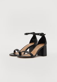 Paire de sandales en cuir noir à talon bloc avec brides à la cheville et une seule bride à l'avant sur un fond blanc uni.