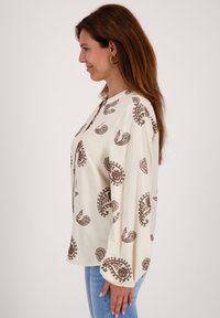 Witte blouse met bruine paisleypatroon, lange mouwen en ronde hals. Gepaard met lichtblauwe jeans.