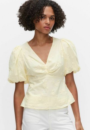 Frau mit lockigem Haar, die eine blassgelbe Bluse mit V-Ausschnitt, Puffärmeln und Blumenstickerei trägt, kombiniert mit einer weißen Hose.