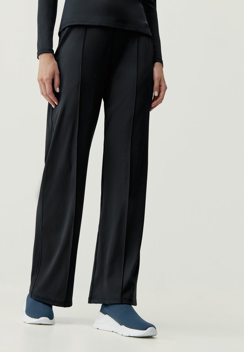 Czarne sportowe spodnie o szerokim kroju, gładkiej fakturze i subtelnych detalach szwów, zestawione z niebieskimi butami wsuwanymi.