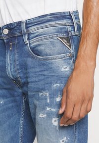 Blue denim jeans featuring a slim fit, faded wash, distressed details, and a black brand tag on the front pocket.