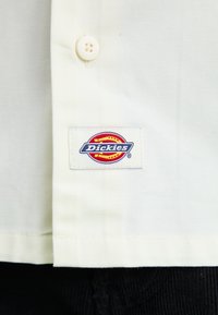 Light beige shirt made of cotton, featuring a button front and a Dickies brand tag. Smooth texture with a simple design.