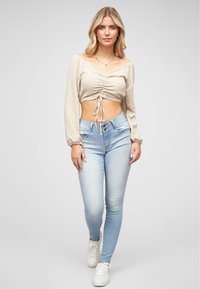 Top beige corto con fronte arricciato e dettaglio con nodo, abbinato a jeans a vita alta blu chiaro e sneakers bianche. Tessuto morbido e design aderente.