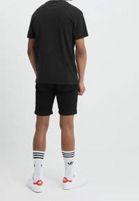 T-shirt nera in cotone e pantaloni corti neri arrotolati, abbinati a calzini bianchi con strisce nere e dettagli rossi, e sneakers bianche.