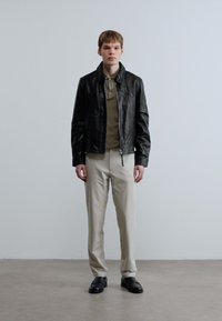 Μαύρο δερμάτινο μπουφάν πάνω από μπεζ πόλο, σετ με ανοιχτό μπεζ παντελόνι και μαύρα παπούτσια. Λείες υφές, εφαρμοστό σχέδιο, απλός εξοπλισμός.