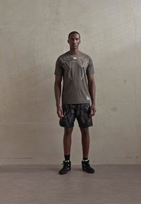 Homme se tenant contre un mur uni, portant un t-shirt graphique olive, un short noir à motifs, des chaussettes noires et des baskets noires.