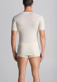 Uomo che indossa una maglietta attillata a maniche corte di colore bianco sporco e pantaloni corti aderenti abbinati, in piedi con la schiena rivolta verso la telecamera su uno sfondo semplice.