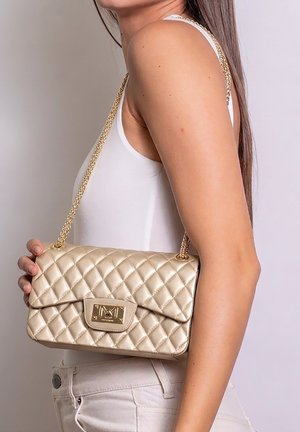 Donna con top e pantaloni bianchi tiene una borsa beige trapuntata con tracolla a catena dorata sulla spalla.