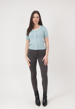 Vrouw staand, draagt een lichtblauwe korte mouw vest, zwarte skinny jeans, zwarte enkellaarsjes, met een tatoeage op de linker onderarm, neutrale achtergrond.