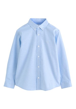 Garçon portant une chemise bleu clair à manches longues boutonnée avec un petit logo de cerf sur la poitrine et un pantalon noir.