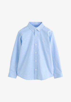 Garçon portant une chemise bleu clair à manches longues boutonnée avec un petit logo de cerf sur la poitrine et un pantalon noir.