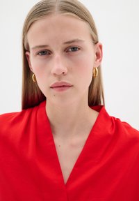 Blusa rossa con scollatura a V, caratterizzata da una texture liscia e un tessuto drappeggiato. Orecchini a cerchio dorati completano l'outfit. Sfondo neutro.