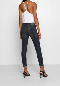 Žena v tmavých úzkých riflích, bílém topu na ramínka a černých vysokých podpatcích, stojící zády k fotoaparátu na jednoduchém pozadí.