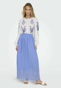 Blusa bianca con ricami floreali blu, abbinata a una lunga gonna blu con dettagli in pizzo all'orlo. Sandali piatti visibili.