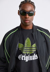 Fiatal férfi, fekete napszemüveget visel, fekete dzseki neon zöld díszítéssel és fekete Adidas Originals pólót nagy zöld logóval.