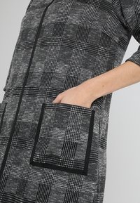 Cappotto lungo nero e grigio con motivo, dotato di una grande tasca frontale, chiusura con zip e maniche a tre quarti per una silhouette strutturata.