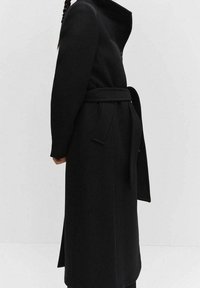 Cappotto in misto lana nero con colletto alto e cintura legata. Caratterizzato da un taglio dritto, maniche lunghe e fessure laterali per facilitare i movimenti.