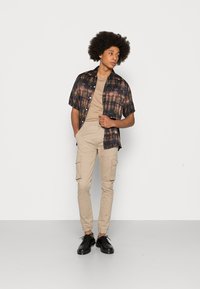 Kortärmad mönstrad skjorta i mörkblått, brunt och beige; beige t-shirt med rund hals; ljusbeige cargobyxor; svarta snörskor.