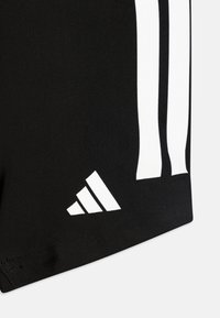 Čierne športové šortky s bielymi akcentovacími pruhmi a trojuholníkovým logom na leme, vyrobené z hladkej, elastickej látky.